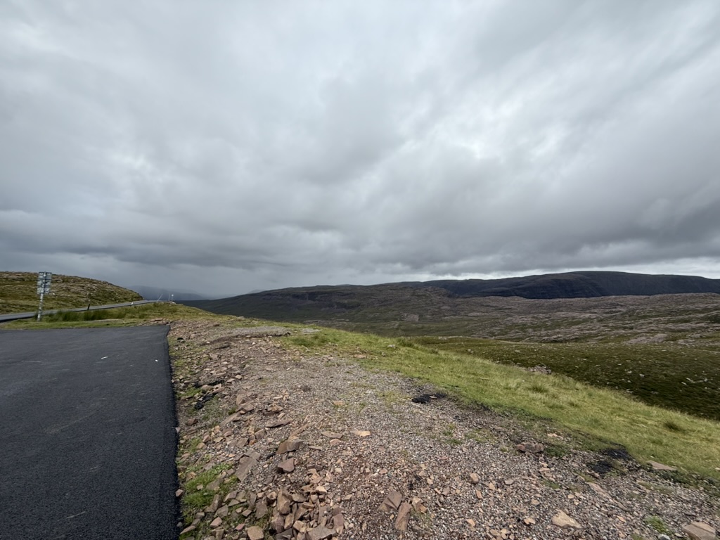 Curving paved road beside rocky, grassy hills under heavy gray clouds on top of Applecross Pass.