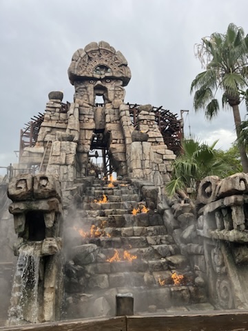 A large stone structure resembling a temple with steps and a waterfall, featuring flames on the stairs and surrounded by palm trees under a cloudy sky.