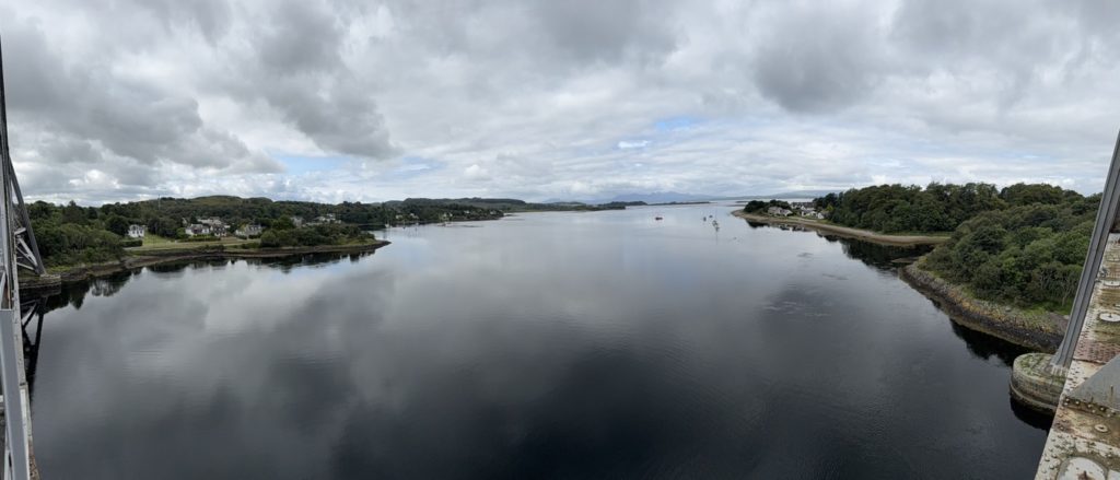 View from Connel Bridge over calm water, houses, and green hills under cloudy sky.