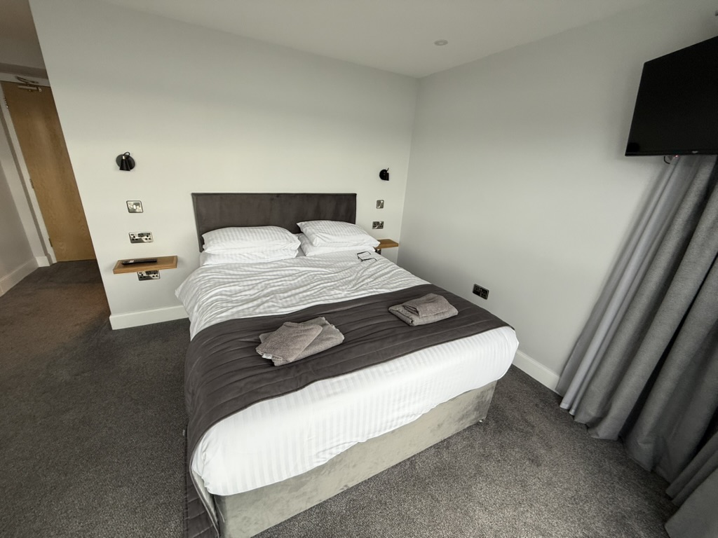 Neatly made hotel bed with white linens, grey throw, and folded towels.