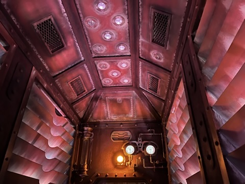 View of a ceiling with a metallic texture, featuring multiple round lights and ventilation grills, illuminated with a pinkish hue.