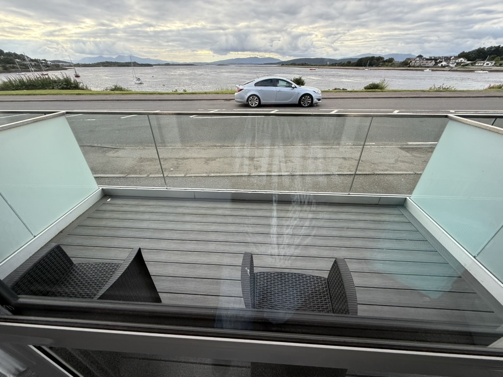 Balcony view of road, water, and anchored sailboats with hills in the distance.