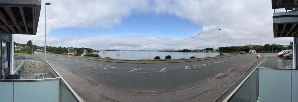 Waterfront view with road, parked cars, boats, and hills under a partly cloudy sky.