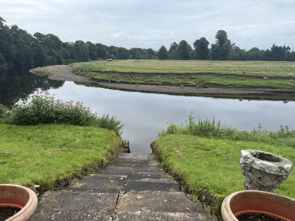 Riverside view at Warrick Hall with stone steps, planters, and grazing sheep across the water.
