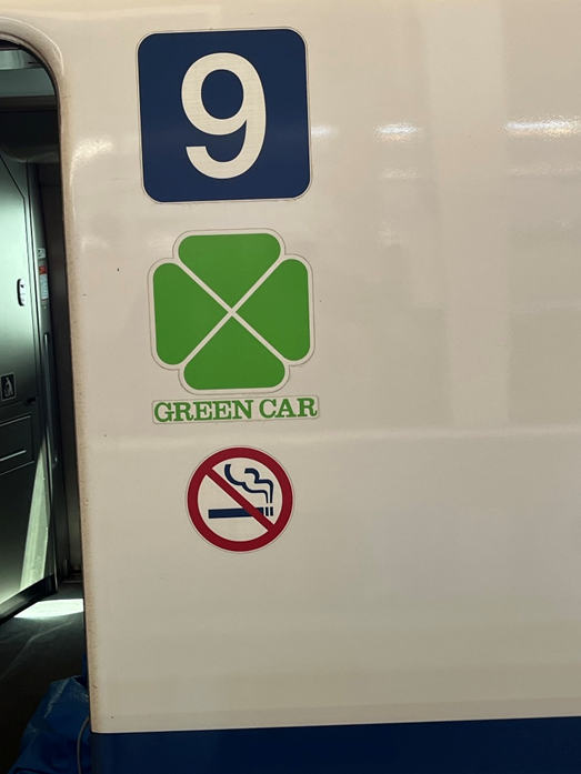 Picture of the side of a train car, with the number 9 blown up in a bit blue box. Underneath that is a green four leaf clover sign with text saying green car. There is also a no smoking sign.
