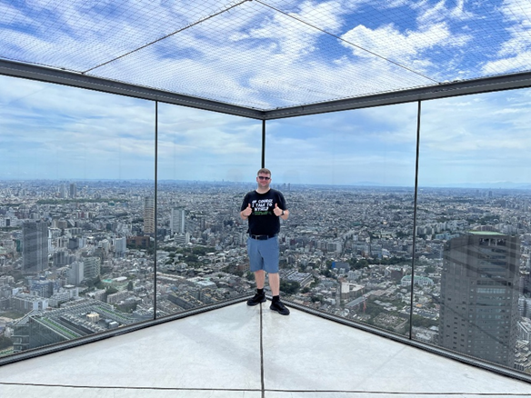 Matthew Jones with his thumbs up and glass behind him with a view out to Tokyo on top of the Shibuya Sky Roof.