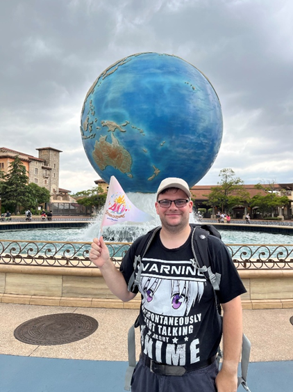 Matthew Jones with the Disneysea globe behind him being held up by a fountain. He is holding a Disney 40th anniversary flag,