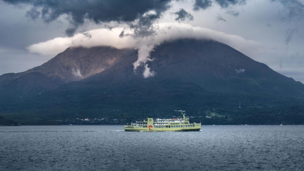 Yellow ferry crossing a bay with a smoking volcanic mountain in the background.